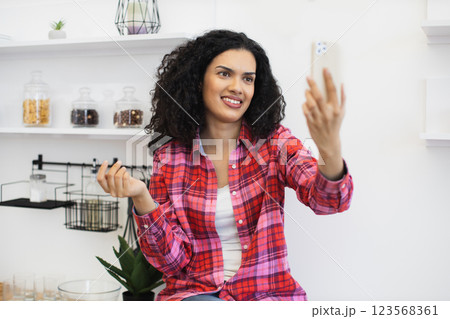 Smiling young African woman in casual plaid shirt taking selfie with smartphone in bright kitchen environment, showcasing expression of joy and friendly personality. 123568361
