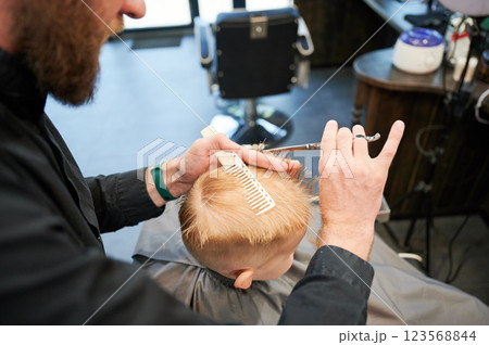 Man barber cutting little boy's hair using comb and scissors. Child getting haircut from adult male, likely barber. Professional hairdresser and cute client at modern barbershop. 123568844