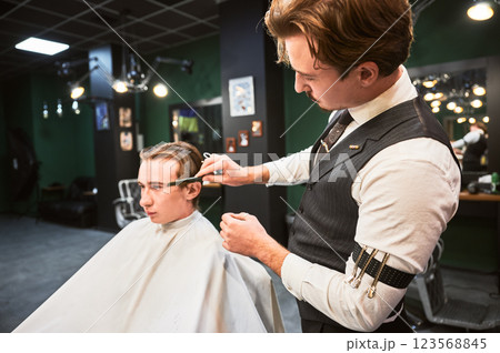 Barber giving haircut to young man in barbershop. Professional barber well-dressed in vest and white shirt with rolled-up sleeves, concentrating on cutting the hair, using comb and scissors. 123568845