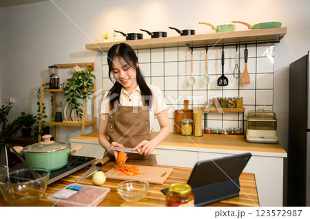 Cheerful young woman wearing a apron slicing carrot while preparing ingredients for homemade spaghetti dish 123572987