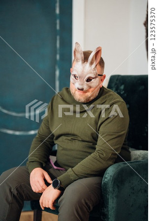 Adult man in green sweater in chair wearing bunny mask. Adult man in green sweater in chair wearing bunny mask. 123575370