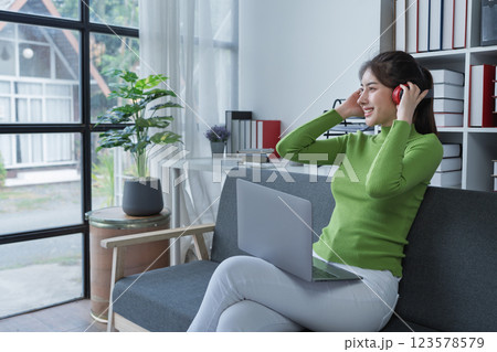 Woman sitting comfortably on a couch with headphones, relaxing to music while using a computer in a cozy setting Woman sitting comfortably on a couch with headphones, relaxing to music while using a computer in a cozy setting 123578579