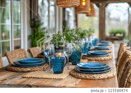 Elegant outdoor dining table set with blue plates, glasses, and wicker chairs in a cozy patio with greenery Elegant outdoor dining table set with blue plates, glasses, and wicker chairs in a cozy patio with greenery 123580974