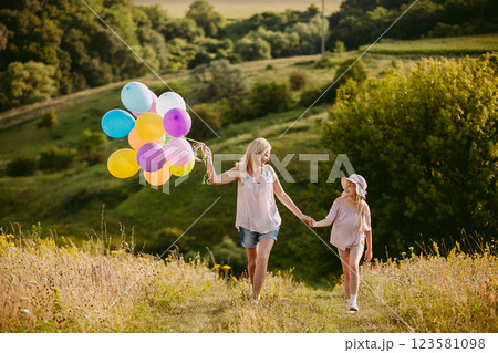 Happy girl with colorful balloons holding hands with a smiling middle-aged woman, walking together in a sunny meadow surrounded by greenery. 123581098