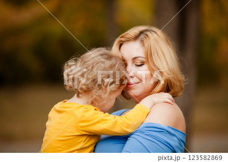 Loving mother holding toddler girl with curly hair in yellow shirt in autumn park 123582869