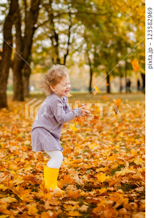 Toddler girl in purple dress, white tights, and yellow rubber boots playing happily with leaves in autumn park 123582969