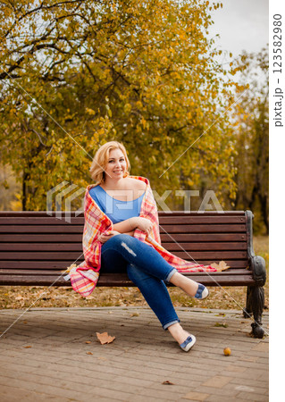 Plus-size blonde woman in jeans and blue shirt sitting on a bench with a blanket on shoulders in autumn park 123582980