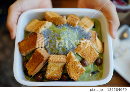 Close up woman hand holding Korean fresh Bingsu sweet ice dessert with green tea ,toast bread and basil seeds topping on white bowl in cafe 123584679