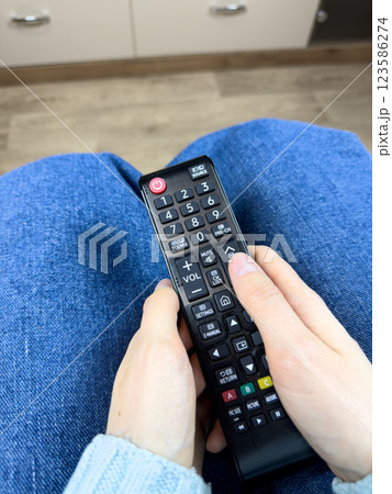 Woman hands hold the remote control on her legs while watching TV. Wireless remote control for controlling sound and changing channels on your TV. Relaxing while watching TV on the sofa 123586274