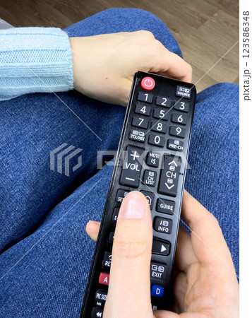 The TV remote control in hands of girl lies on her legs while watching an interesting television program. Control your TV at home using remote controller, vertical photo of TV controller 123586348