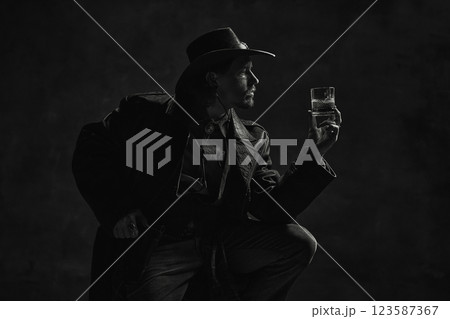 Black and white portrait of man admiring whiskey glass, dressed in leather coat and hat against dark, dramatic background. 123587367