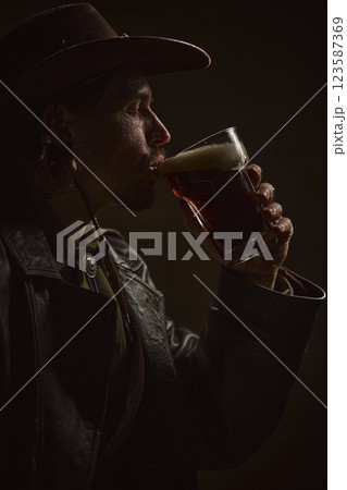 Close-up of man, cowboy sipping dark beer, eyes closed, savoring rich taste, dressed in western attire with hat and leather coat 123587369