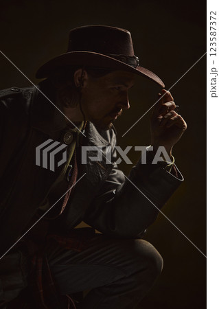 Portrait of brutal thoughtful man, cowboy dressed in leather coat and hat exuding silent strength and wisdom against dark background. Portrait of brutal thoughtful man, cowboy dressed in leather coat and hat exuding silent strength and wisdom against dark background. 123587372