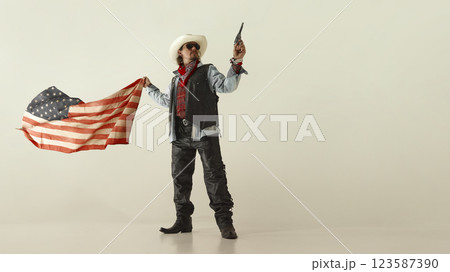 Powerful cowboy in white hat and leather vest, pointing revolver forward, gripping American flag, wearing sunglasses and boots. 123587390