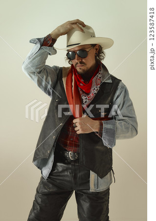Cool man, cowboy adjusting white hat, wearing sunglasses, red bandana, and black leather vest, gripping American flag scarf with serious look. 123587418