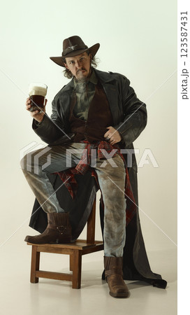 Brutal man dressed as cowboy sitting on chair, smoking and holding glass of dark foam beer. 123587431