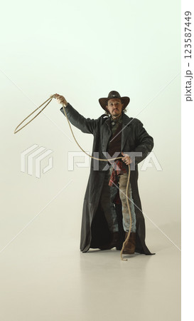 Full-length portrait of brutal man in vintage style country clothes, swinging lasso, ready to throw against light studio background 123587449