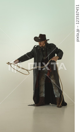 Skilled cowboy swings lasso with sharp focus, gripping rope firmly, dressed in leather coat and wide-brimmed hat against light studio background Skilled cowboy swings lasso with sharp focus, gripping rope firmly, dressed in leather coat and wide-brimmed hat against light studio background 123587451