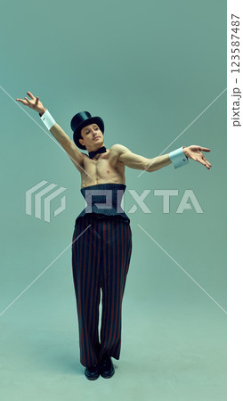 Enigmatic figure in structured corset suit and sleek hat raising cane dramatically against muted green studio background. Enigmatic figure in structured corset suit and sleek hat raising cane dramatically against muted green studio background. 123587487