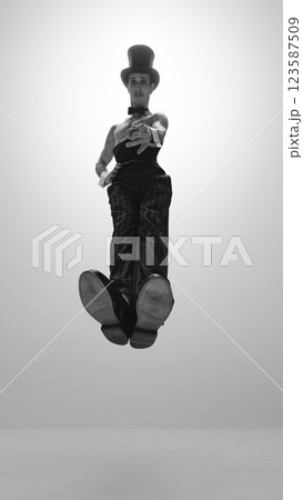 Graceful man wearing in top hat and bow tie levitating while reaching forward against white studio background. Black and white photo. Graceful man wearing in top hat and bow tie levitating while reaching forward against white studio background. Black and white photo. 123587509