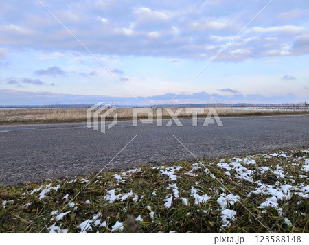 A serene rural road runs through fields under a bright blue sky tinged with purple. The spring road through the field. Early spring, green grass under the snow along the road. A serene rural road runs through fields under a bright blue sky tinged with purple. The spring road through the field. Early spring, green grass under the snow along the road. 123588148
