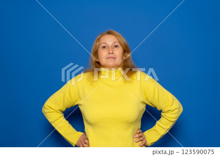 Caucasian woman in her 40s wearing yellow sweater posing happy with arms on hips and looking at camera over blue background with copy space. Confident woman is happy and feeling relaxed Caucasian woman in her 40s wearing yellow sweater posing happy with arms on hips and looking at camera over blue background with copy space. Confident woman is happy and feeling relaxed 123590075