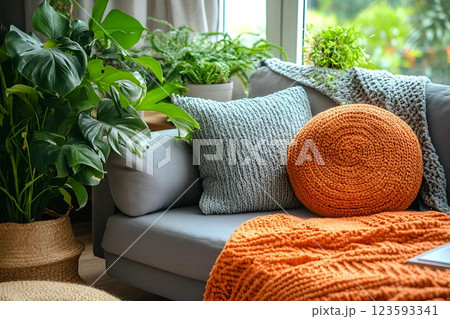 Cozy living room corner with a gray couch, knitted pillows, and an orange throw blanket. Surrounded by lush green plants 123593341