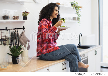 African woman in twenties, wearing plaid shirt and jeans, eating salad indoors on kitchen counter, enjoying fresh food and modern lifestyle, surrounded by a clean and organized interior. African woman in twenties, wearing plaid shirt and jeans, eating salad indoors on kitchen counter, enjoying fresh food and modern lifestyle, surrounded by a clean and organized interior. 123599657
