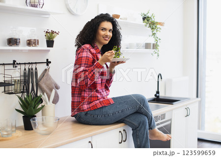 African woman in twenties, wearing plaid shirt and jeans, eating salad indoors on kitchen counter, enjoying fresh food and modern lifestyle, surrounded by a clean and organized interior. African woman in twenties, wearing plaid shirt and jeans, eating salad indoors on kitchen counter, enjoying fresh food and modern lifestyle, surrounded by a clean and organized interior. 123599659