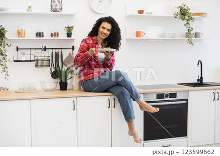 Joyful young African woman in a casual kitchen setting sitting on counter holding plate. Bright and modern interior with a cozy vibe showcasing home lifestyle and meal enjoyment. Joyful young African woman in a casual kitchen setting sitting on counter holding plate. Bright and modern interior with a cozy vibe showcasing home lifestyle and meal enjoyment. 123599662