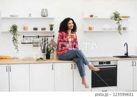 African American woman in casual attire sitting on kitchen island holding coffee cup, morning relaxed mood in modern kitchen interior with plants, utensils, open shelves with cups. African American woman in casual attire sitting on kitchen island holding coffee cup, morning relaxed mood in modern kitchen interior with plants, utensils, open shelves with cups. 123599681
