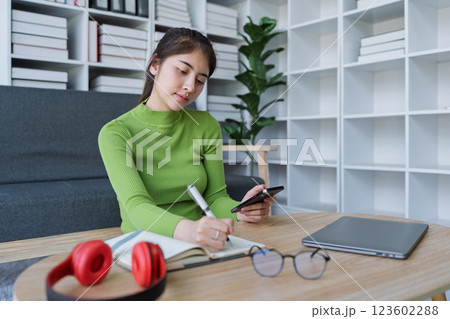 Woman sitting at a table with a laptop and cellphone using smartphone to take notes and work in a cozy home 123602288