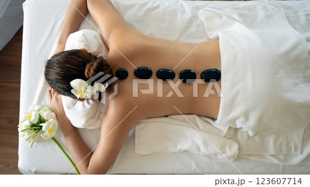 Young woman receiving hot stone therapy lying face down on massage table with black basalt stones aligned on her bare back, adorned with white flowers in hair Young woman receiving hot stone therapy lying face down on massage table with black basalt stones aligned on her bare back, adorned with white flowers in hair 123607714