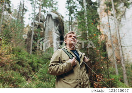 A tourist with a backpack stands against the background of a mountains and forest. Young 30s blonde man hiking in nature. Travel, adventure. Concept of an active lifestyle. High quality photo. 123610097