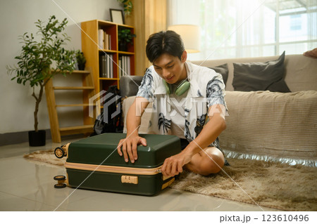 Happy young man sitting on the floor closing his suitcase, preparing for a summer trip Happy young man sitting on the floor closing his suitcase, preparing for a summer trip 123610496