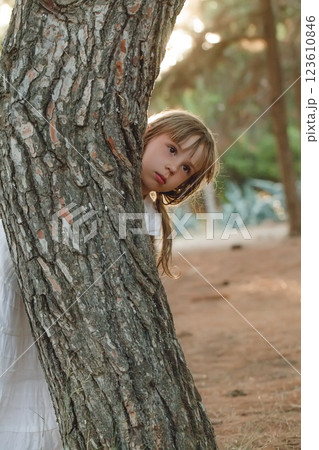 Child girl in white clothes peeking out from behind the tree in the park Child girl in white clothes peeking out from behind the tree in the park 123610846
