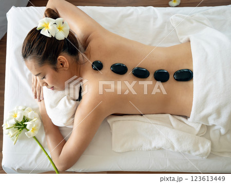 Young woman receiving hot stone massage in spa, lying on massage bed with black basalt stones placed along her spine, surrounded by white plumeria flowers 123613449