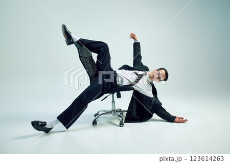 Young man dressed formal, black attire lying in office chair with raising arms and legs, with questioning expression against white studio background. 123614263
