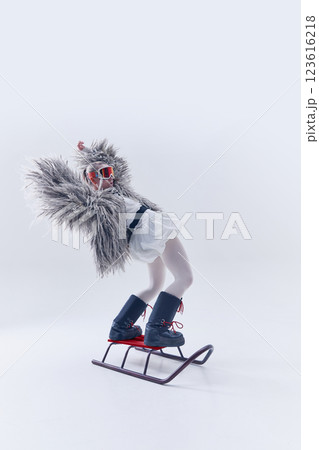 Young woman leaning back on sled in oversized shaggy fur coat, ski goggles, and moon boots against white studio background. 123616218