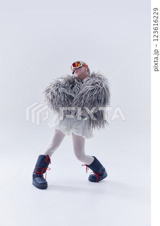 Young woman striking stylish pose in oversized shaggy fur coat, ski goggles, and moon boots against white studio background Young woman striking stylish pose in oversized shaggy fur coat, ski goggles, and moon boots against white studio background 123616229
