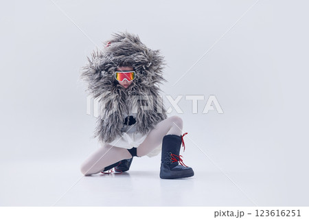 Young woman posing in oversized shaggy fur coat, ski goggles, and moon boots against white studio background. Young woman posing in oversized shaggy fur coat, ski goggles, and moon boots against white studio background. 123616251