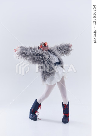 Young woman extending arms in oversized shaggy fur coat, ski goggles, and moon boots against white studio background. 123616254
