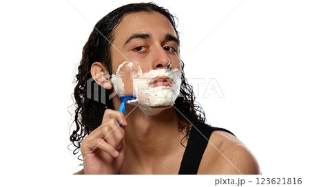 Young man with curly wet hair shaving face with blue razor, thick foam covering skin against white studio background. 123621816