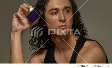 Stylish young guy with wet curly hair holding luxury skincare product, cream near face against studio background 123621905