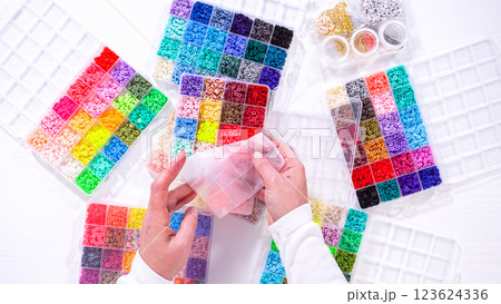 Flat lay. Woman hands gracefully poised over a collection of beads, sorted by color in transparent organizers. The array of beads spans a vibrant spectrum, from deep purples to bright oranges 123624336
