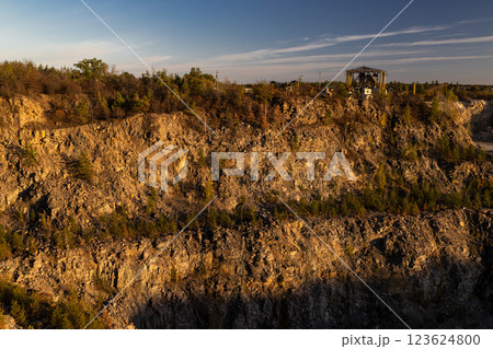 A rocky cliff with sparse trees and dry bushes under warm sunlight. At the top, an abandoned industrial structure stands among autumn foliage, creating a contrast between nature and human-made ruins. 123624800