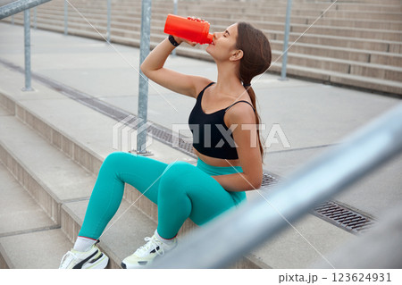Young Woman Drinks Water While Resting on Stairs After Workout in Urban Environment During Daylight Young Woman Drinks Water While Resting on Stairs After Workout in Urban Environment During Daylight 123624931