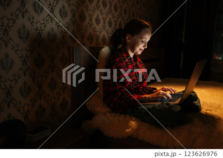 Side view of focused schoolgirl studying on laptop sitting on bed at night, face illuminated by screen soft light in darkened bedroom, creating cozy atmosphere during peaceful nighttime. 123626976