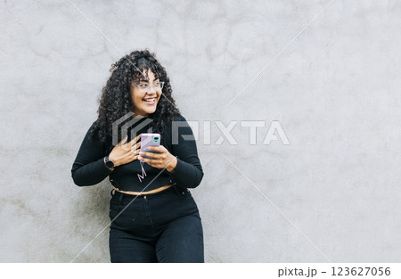 young latin woman happy with her cell phone leaning against a wall. A young latin woman laughing with her cell phone leaning against a wall, Pretty girl happy with her cell phone young latin woman happy with her cell phone leaning against a wall. A young latin woman laughing with her cell phone leaning against a wall, Pretty girl happy with her cell phone 123627056