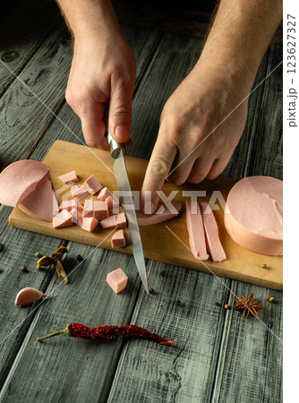 Hands expertly slice a slab of sausage for lunch, creating neat cubes and strips on a rustic wooden cutting board surrounded by vibrant spices and chili peppers 123627327